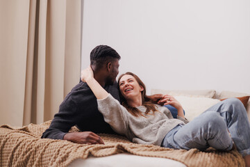 Smiling woman talking to african american boyfriend while relaxing on bed at home 