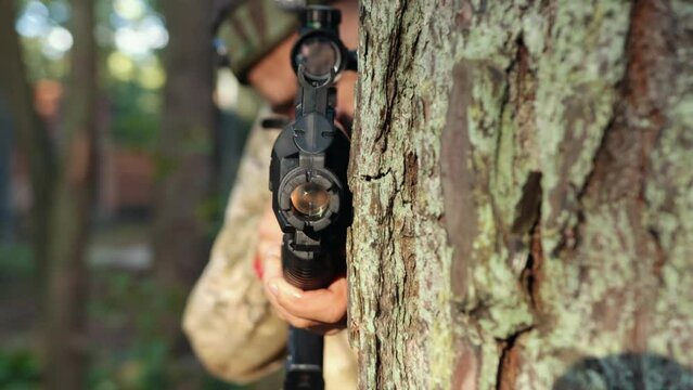 Camouflage shooter, forest skirmish, aimed shot. In forested setting, man adorned in camo gear with marker on his head is positioned against tree, skillfully targeting with Laser tag gun