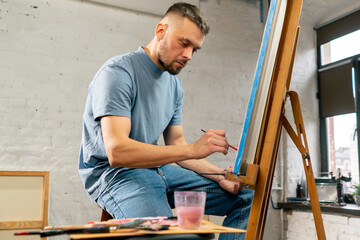 Fototapeta na wymiar young artist in a blue t-shirt in an art studio working on painting while sitting