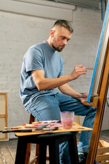 Naklejka na ściany i meble young artist in a blue t-shirt in an art studio working on painting while sitting