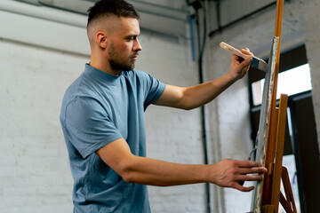 in an art workshop an artist in a blue T-shirt makes broad strokes on the canvas with wide and flat brush