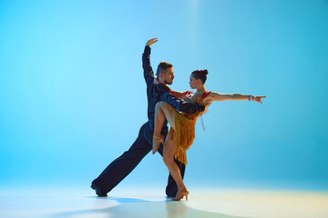Young man and woman in elegant stage costumes performing samba, dancing against gradient blue background in neon light. Concept of dance class, hobby, art, dance school, talent