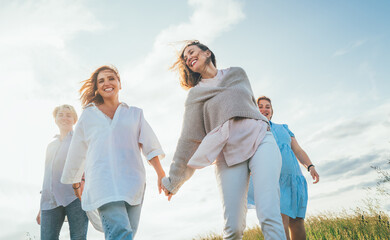 Portrait of four cheerful smiling women holding hand in hand walking by a high green grass meadow....