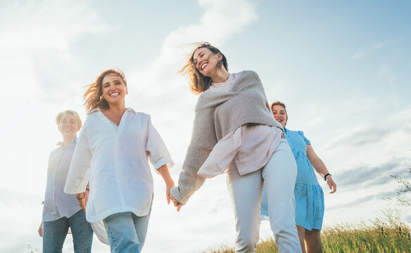 Portrait of four cheerful smiling women holding hand in hand walking by a high green grass meadow. They looking at the camera. Woman's friendship, relations, and happiness concept image.