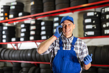 Positive auto mechanic talking on a mobile phone against the background of car tires in a car...