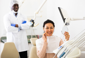 Satisfied asian woman looking in the mirror at the cured teeth at the dentist