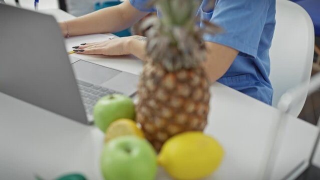 A focused woman in scrubs uses a laptop in a clinic surrounded by fresh fruit and a measuring tape, depicting a nutritionist at work.