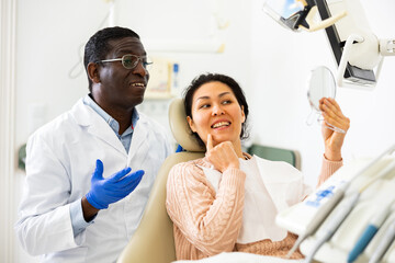 Positive asian woman and african-american man dentist looking at teeth through mirror after dental...
