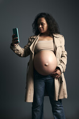 Pregnant woman with naked belly, in stylish beige trench, using smart mobile phone, isolated over gray studio background