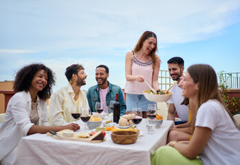A group of people is gathered around a table, sharing food and wine while smiling and enjoying the leisurely event. The hostess serves food to her guests. Friends lunch together on a spring summer day