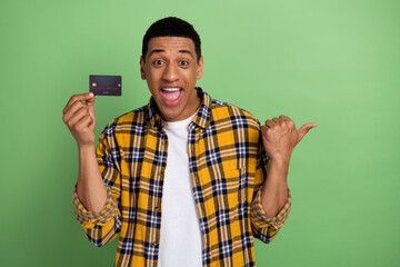 Photo of young crazy guy wearing plaid yellow shirt holding black bank card advertise finger mockup terms isolated on green color background