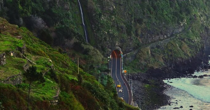 Aerial view scenic road along the Atlantic Ocean with a tunnel for cars above which a waterfall cascades down the mountain, located on the island of Madeira, on the north side of the island, Portugal