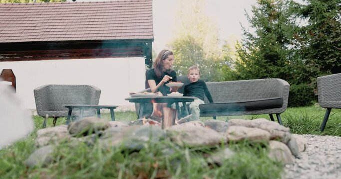 Nourishing moments blossom as a mother and child share food in the backyard patio, by the flickering flames of the fire pit