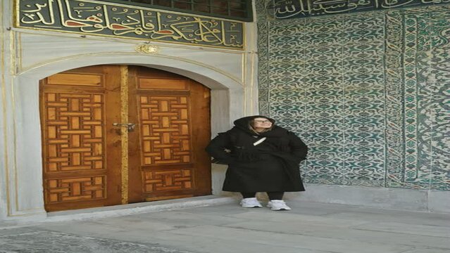 A smiling woman in a black outfit leaning against an ornate wall with islamic calligraphy at the topkapi palace in istanbul.