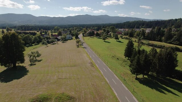 Aerial view of a countryside area in Slovenia, there is a small village and a road that cuts through the fields. In the background there is a pine forest and the Balkan mountains.