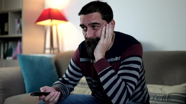 Bearded man holding remote control watching tv sitting on sofa with boring expression at home.