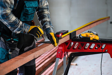 Fototapeta premium a worker in fitters and gloves with a tool belt measures wood using a tape measure and a right-angled triangle, blurred background