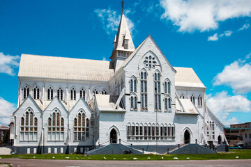 A tall white wooden cathedral with a spire and elongated windows on a sunny day against a blue sky with clouds, Guyana. World tourism, architecture.