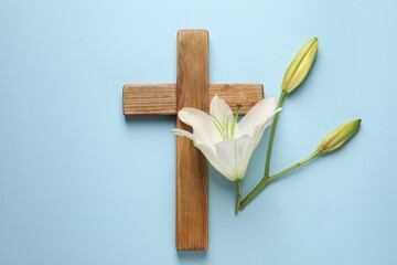 Wooden cross and lily flowers on light blue background, top view. Easter attributes