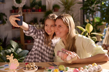 Cheerful Caucasian mother and daughter sitting at table in workshop taking selfie on smartphone while decorating eggs for Easter