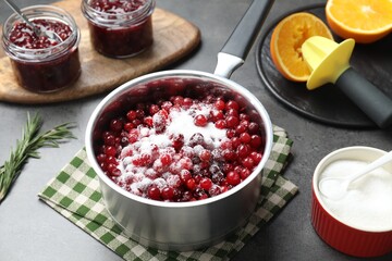 Making cranberry sauce. Fresh cranberries with sugar in saucepan and ingredients on gray table, closeup
