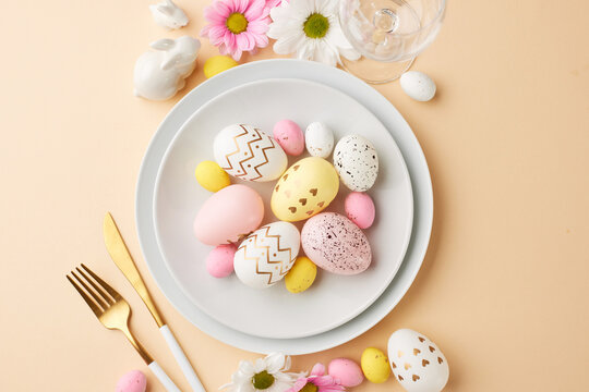 Easter gathering: a table set for celebration. Top view shot of plate with gold cutlery and decorated Easter eggs, white and pink daisies, rabbit figure and wine glass on a pastel peach background