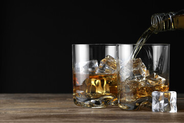 Pouring whiskey into glass with ice cubes at wooden table against black background, space for text
