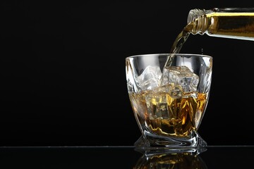 Pouring whiskey from bottle into glass with ice cubes at table against black background, space for text