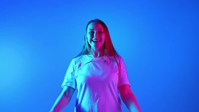 Woman with freckles and ginger hair laughing looking at camera and clenching fists against gradient blue studio background in neon light. Concept of beauty, fashion, self-expression, human emotions.