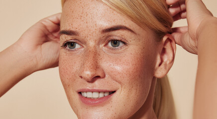 Close up highly detailed portrait of a young smiling woman with freckles. Cropped image of a beautiful blond female adjusting hair against a beige backdrop.