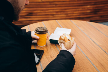 A man eats a burger and drinks coffee while sitting at a wooden table outside in a fast food cafe....