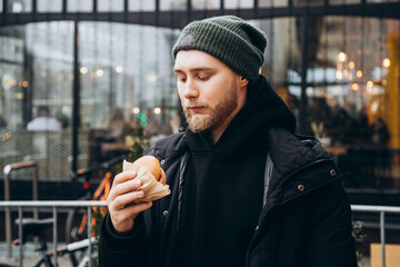 Attractive bearded young man wearing a jacket and hat with a burger outside. A man eats fast food...