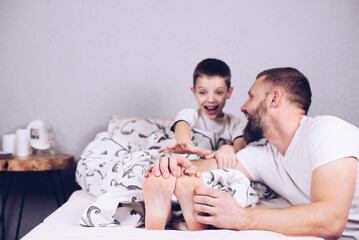 A child with bare feet is lying in bed. Dad tickles his son. Tickling the heels. Cheerful and...