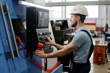 Side view of young bearded Caucasian man wearing hardhat operating modern CNC machine