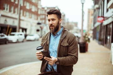 Man with beard holding coffee and smartphone on city street