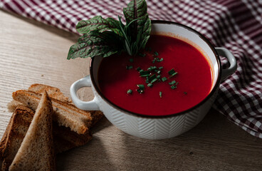 beetroot tomatoes soup in white bowl with handles served with crunchy triangle toasted bread, green beet leaves, green garlic, rustic cloth and wooden background