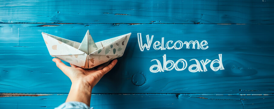 Hand presenting a paper boat with the message Welcome aboard against a blue background, symbolizing new beginnings and joining a team or company