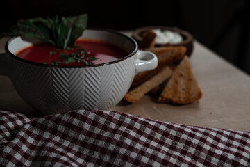 beetroot tomatoes soup in white bowl with handles served with crunchy triangle toasted bread, green beet leaves, green garlic, rustic cloth and wooden background