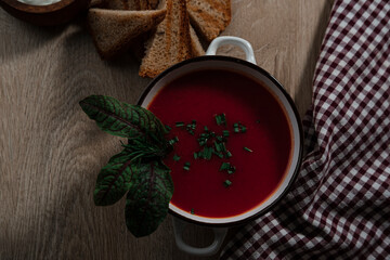 beetroot tomatoes soup in white bowl with handles served with crunchy triangle toasted bread, green beet leaves, green garlic, rustic cloth and wooden background