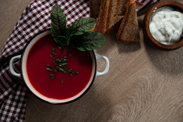 beetroot tomatoes soup in white bowl with handles served with crunchy triangle toasted bread, green beet leaves, green garlic, rustic cloth and wooden background