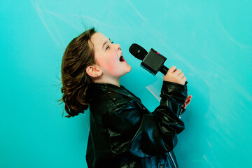 A young girl in a black leather jacket belts out a song into a microphone with a vibrant turquoise background illuminating her