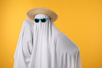 Person in ghost costume, sunglasses and straw hat on yellow background, space for text