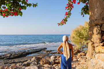 Beautiful woman in Greek dress and arms akimbo enjoying a sunrise by the sea and observing a...