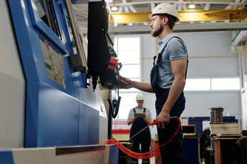 Low angle medium long side view of bearded Caucasian man wearing uniform with hardhat using CNC...