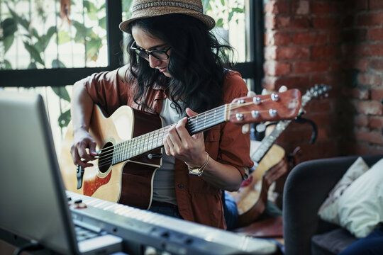 Generative AI illustration of woman sits in a home music studio, playing a guitar while interacting with a laptop