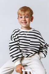 Blonde little kid in striped sweater and white trousers sitting posing at studio