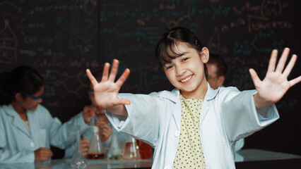 Cute asian scientist waving hand to camera while diverse group doing experiment. Young schoolgirl standing at blackboard with chemical theory written while wearing lab coat at laboratory. Edification.