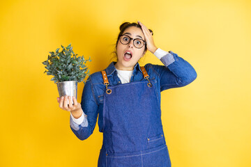 Young caucasian gardener woman holding a plant isolated on yellow background putting one hand on...