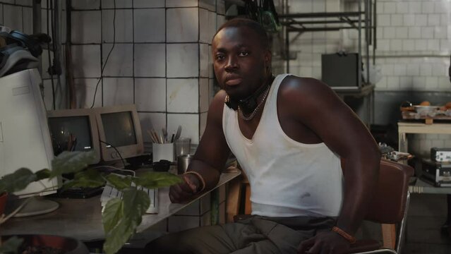Portrait of young Black man sitting at desk with old computer and posing for camera in post apocalypse bunker