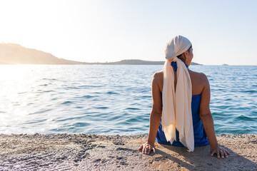 Beautiful close up of woman with bare back and headscarf by the sea sitting on a jetty at dawn in...